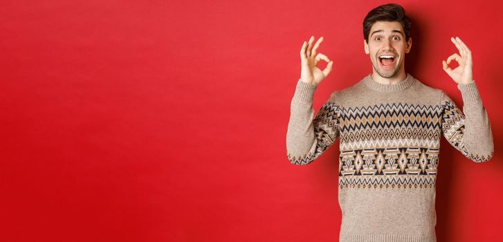 Concept of christmas celebration, winter holidays and lifestyle. Portrait of handsome man in sweater, looking amazed and showing okay signs, guarantee or recommend something good, red background.