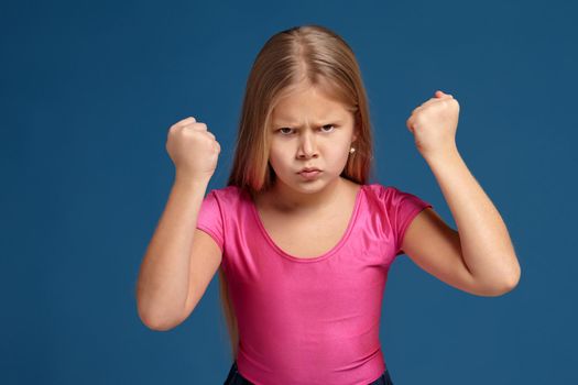Portrait of emotional blonde little girl on blue background. in pink shirt, acting like anger unhappy person, treatens with her fists