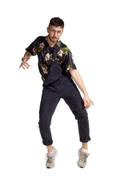 Full-length portrait of a handsome male in glasses, black jumpsuit, colorful t-shirt and gray sneakers fooling around in studio. Indoor photo of a man dancing isolated on white background. Music and imagination concept.