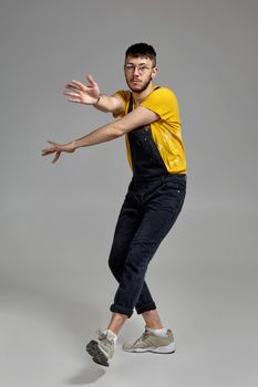 Full-length portrait of an athletic dancer in glasses, black jumpsuit, yellow t-shirt and gray sneakers fooling around in studio. Indoor photo of a man dancing on a gray background. Music and imagination.