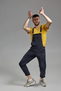 Full-length portrait of a sporty guy in glasses, black jumpsuit, yellow t-shirt and gray sneakers fooling around in studio. Indoor photo of a man dancing raising his hands on a gray background. Music and imagination.