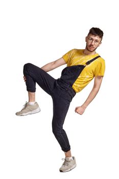 Full-length portrait of a good-looking dancer in glasses, black jumpsuit, yellow t-shirt and gray sneakers fooling around in studio. Indoor photo of a man making dancing elements raising his leg isolated on white background. Music and imagination.