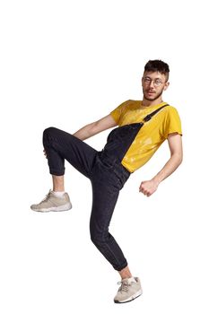 Full-length portrait of a good-looking man in glasses, black jumpsuit, yellow t-shirt and gray sneakers fooling around in studio. Indoor photo of a man making dancing elements raising his leg isolated on white background. Music and imagination.