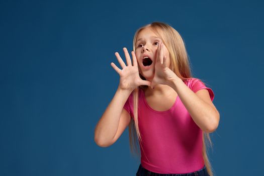 Portrait of adorable blonde little girl on blue background. in pink shirt shouting, trying to call someone