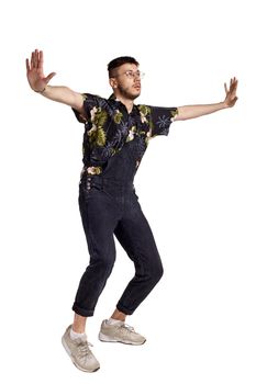 Full-length portrait of a good-looking dancer in glasses, black jumpsuit, colorful t-shirt and gray sneakers fooling around in studio. Indoor photo of a man dancing sideways isolated on white background. Music and imagination.