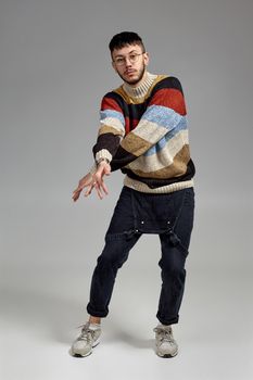 Full-length portrait of an athletic person in glasses, black jumpsuit, multi-colored sweater and gray sneakers fooling around in studio. Indoor photo of a male dancing on a gray background. Music and imagination. Copy space.