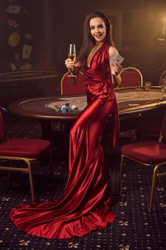 Young brunette female in a long burgundy atlas dress, with two aces and a glass of champagne in her hands is posing against a poker table in vip casino. Passion, cards, chips, alcohol drinks, win, gambling - it is a best entertainment.