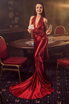 Young brunette maiden in a long burgundy atlas dress, with two aces and a glass of champagne in her hands is smiling and posing against a poker table in vip casino. Passion, cards, chips, alcohol drinks, win, gambling - it is a best entertainment.