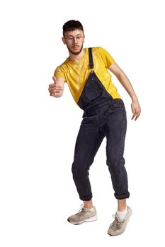 Full-length portrait of a stylish man in glasses, black jumpsuit, yellow t-shirt and gray sneakers fooling around in studio. Indoor photo of a man making dancing elements isolated on white background. Music and imagination.