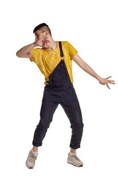 Full-length portrait of an elegant male in glasses, black jumpsuit, yellow t-shirt and gray sneakers fooling around in studio. Indoor photo of a man making dancing elements isolated on white background. Music and imagination.