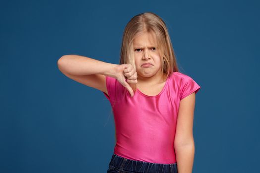 Close-up emotional portrait of caucasian little girl in pink shirt on blue background, making dislike, thumbs down gesture, looks unhappy