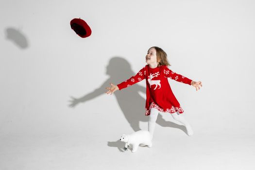 Studio portrait of cheerful little girl in red dress with reindeer and snowflakes throwing beret and running after it. White bear toy on the floor.