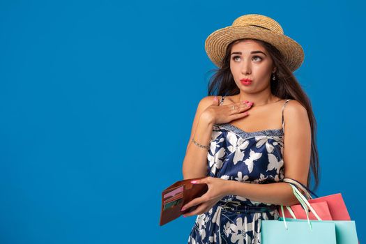 Portrait of unhappy young woman looking in her wallet in shopping center, spent too much, not enough cash, lost money, broke. Studio shot on blue background. Copy space