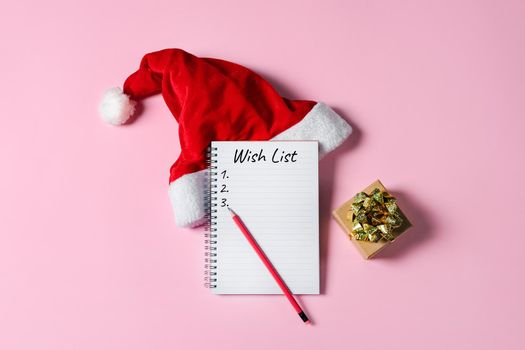 New Year or Christmas wish list concept. Notepad, Santa Claus hat and gift box isolated on pink background. Holiday banner with copy space.