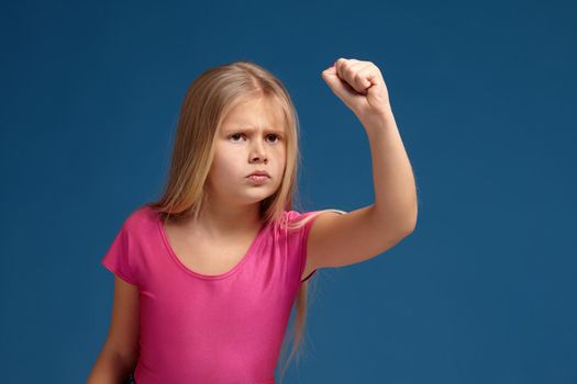 Portrait of emotional blonde little girl on blue background. in pink shirt, acting like anger unhappy person