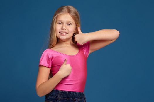Close-up emotional portrait of caucasian little girl in pink shirt on blue background, making thumbs up gesture, looks happy