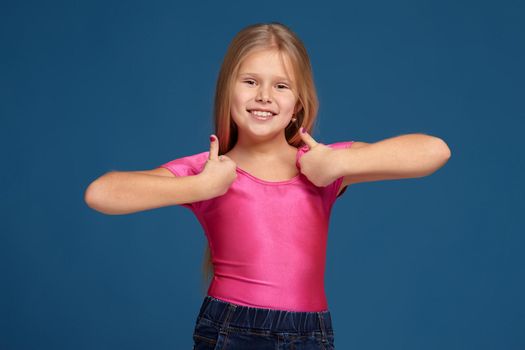 Close-up emotional portrait of caucasian little girl in pink shirt on blue background, making thumbs up gesture, looks happy