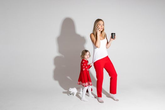 Busy mom talking via cell and drinking coffee while her daughter in red dress with Christmas pattern pulling her top demanding attention.