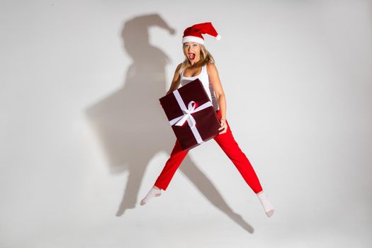 Stock photo of jumping cheerful blonde woman in red Santa hat and red trousers with a wrapped Christmas gift with open mouth.