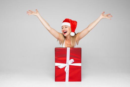 Merry young lady in Santa hat putting hands up and smiling widely while popping out of a gift box. Copy space. Holiday concept