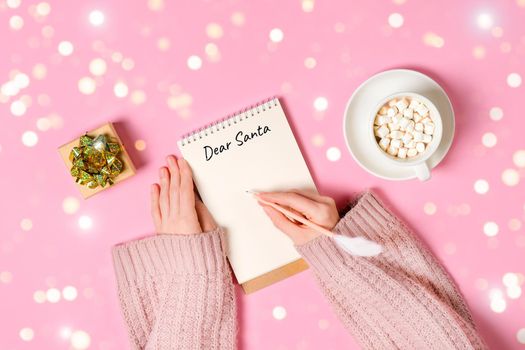 Letter to Dear Santa. Hand holding pink pen decorated with feather and writing in Notepad letter to Santa Claus on pink background with cup of hot chocolate with marshmallows, a gift, golden tinsel.