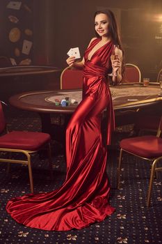 Young brunette lady in a long burgundy atlas dress, with two aces and a glass of champagne in her hands is posing near a poker table in vip casino. Passion, cards, chips, alcohol drinks, win, gambling - it is a best entertainment.