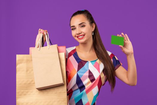 Attractive woman 20s in dress holding shopping packages and showing green credit card isolated over purple background. Studio shot. Copy space