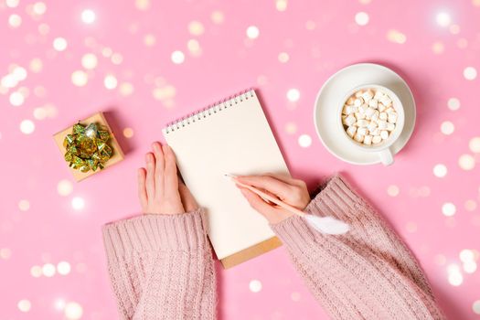 New year, Christmas or holiday wish list concept. Notepad, golden tinsel, and a cup of hot chocolate with marshmallows and female's hands writing isolated on pink background. Banner with copy space