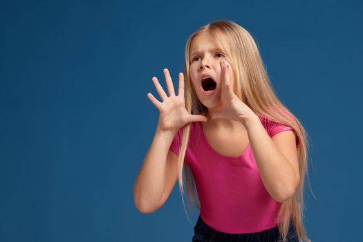 Portrait of adorable blonde little girl on blue background. in pink shirt shouting, trying to call someone