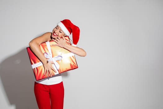 Stock photo of beautiful delighted woman in Santa hat with eyes closed hugging red wrapped Christmas gift with white bow.