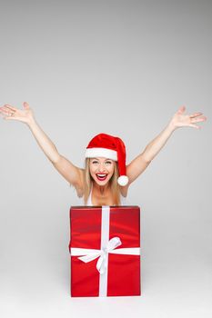 Merry young lady in Santa hat putting hands up and smiling widely while popping out of a gift box. Copy space. Holiday concept