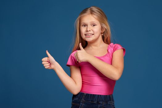Close-up emotional portrait of caucasian little girl in pink shirt on blue background, making thumbs up gesture, looks happy