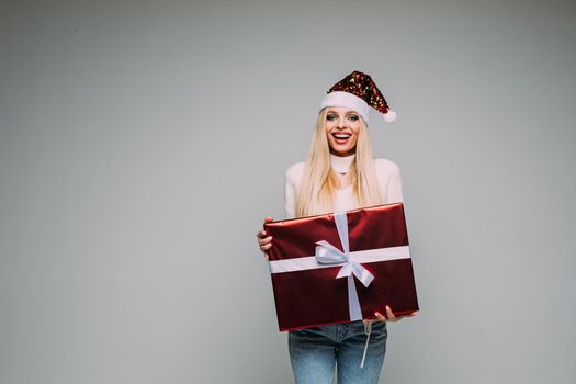 Happy Caucasian woman in white sweater and Santa hat holding gift box and looking at camera. Holiday concept