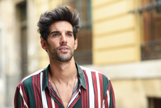 Attractive young man with dark hair and modern hairstyle wearing casual clothes in urban background.