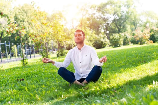 Young man meditates in lotus pose on green grass. Handsome man in casual wear practicing of yoga with closed eyes. Training and meditation outdoor at summer day. Healthy lifestyle and relaxation.