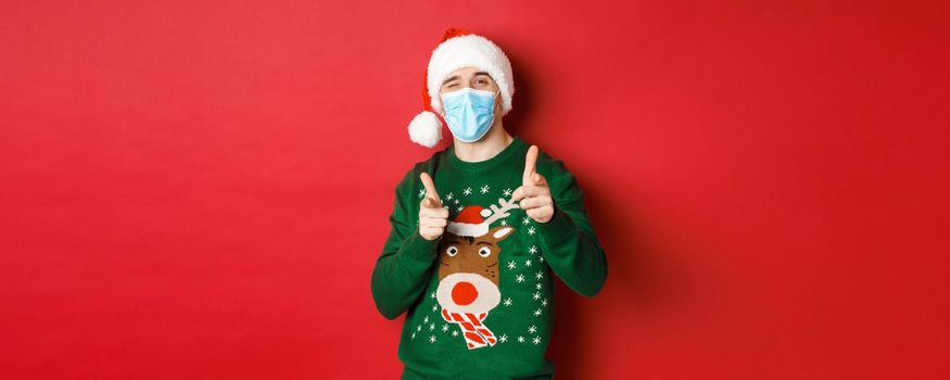 Concept of new year, covid-19 and social distancing. Cheerful man in christmas sweater, medical mask and santa hat, pointing fingers at camera, wishing happy holidays, standing over red background.