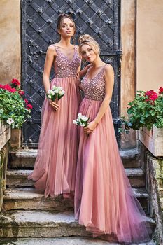 Beautiful bridesmaids in gorgeous elegant stylish red pink violet floor length v neck chiffon gown dress decorated with sequins sparkles and rhinestones holding flowers. Wedding day in old beautiful European city.