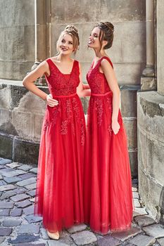 Two beautiful bridesmaids blonde and brunette ladies wearing elegant full length red chiffon bridesmaid dress with lace and holding flower bouquets. European old town location for wedding day.