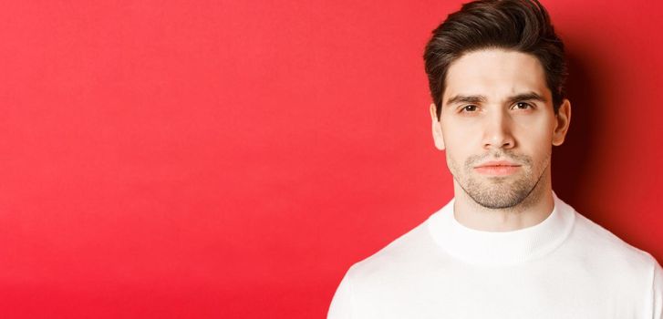 Close-up of thoughtful, serious-looking man in white sweater, standing over red background.