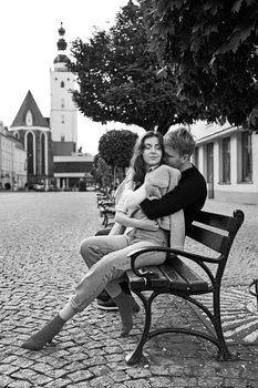 Beautiful young caucasian couple cuddling on a bench in a European town. Romatnic date and love concept.
