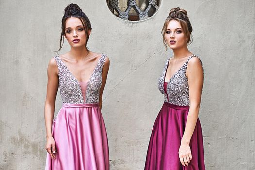 Two beautiful bridesmaids girls blonde and brunette ladies wearing elegant full length purple violet lilac lavender satin bridesmaid dress with silver sequined camisole top. European old town location for wedding day.