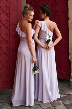 Two beautiful bridesmaids girls blonde and brunette ladies wearing full length lavender tulle one shoulder bridesmaid dress and holding flower bouquets. European old town location for wedding day.