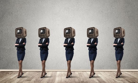Business women in suits with old TV instead of their heads keeping arms crossed while standing in a row in empty room with gray wall on background.