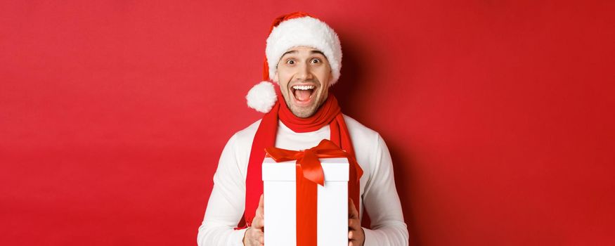 Concept of winter holidays, christmas and lifestyle. Close-up of excited handsome man in santa jat and scarf, looking amazed and receiving new year present, standing over red background.