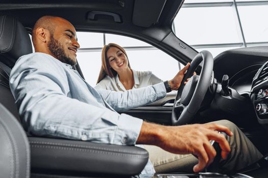 Young attractive caucasian woman salesperson in car showroom showing a car to her male African American client