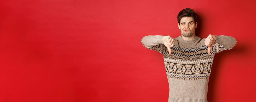 Portrait of skeptical and disappointed handsome man in christmas sweater, dislike new year party, showing thumbs-down, express disapproval, standing over red background.