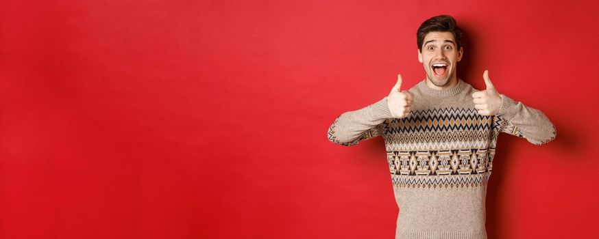 Portrait of cheerful man in christmas sweater like something good, showing thumbs-up and smiling amazed, praise new year party, standing over red background.