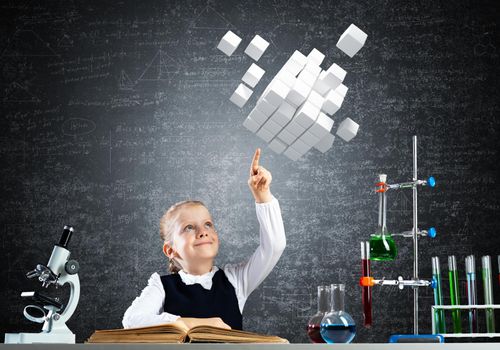 Little girl scientist with microscope on chalkboard background. Research and discovery. Elementary science class in school. Schoolgirl in schoolwear sits at desk and finger pointing on 3d cubes