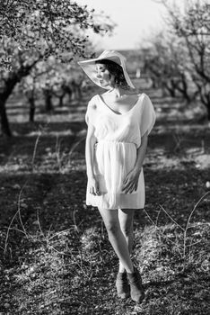 Portrait of young woman in the flowered field in the spring time. Almond flowers blossoms. Girl wearing white dress and pink sun hat