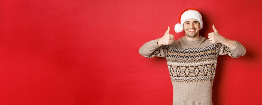 Portrait of attractive happy man in santa hat and sweater, showing thumbs-up in approval and smiling, wishing merry christmas, standing over red background.